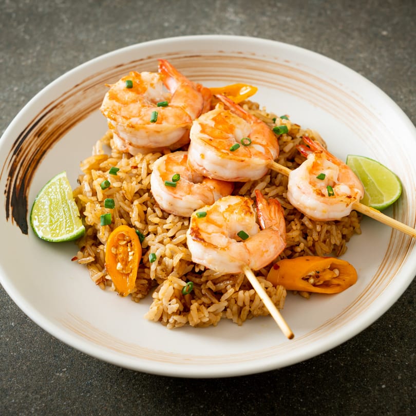 Shrimp skewers with fried rice, garnished with lime wedges and sliced peppers on a white plate.