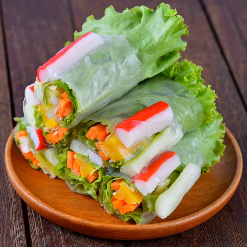 Vietnamese spring rolls with crab sticks, lettuce, and vegetables on a wooden plate.